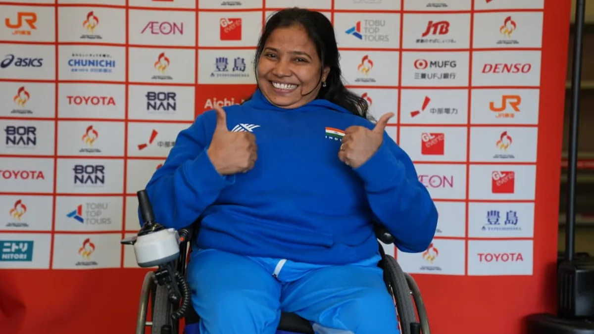 India's Suvarna Raj in a blue India tracksuit giving two thumbs up from her wheelchair in front of the Aichi-Nagoya 2026 Asian Para Games sponsor wall